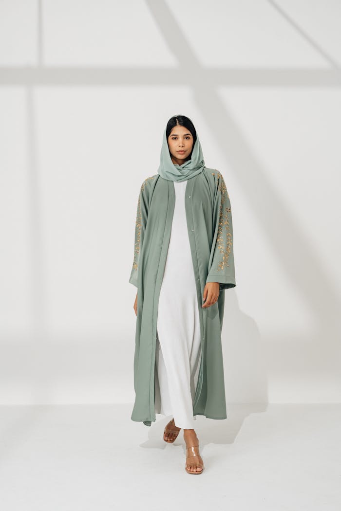 Graceful female in green embroidered abaya and hijab standing in studio light setting.