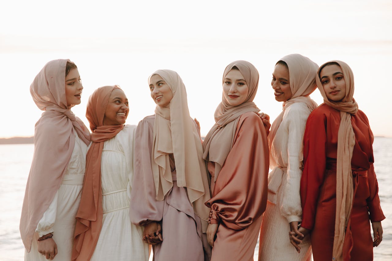 A group of women in fashionable hijabs and dresses, smiling by the sea at sunset.
